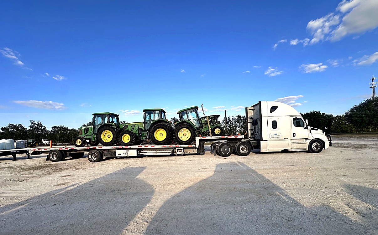 Oneway Group stepdeck loaded with green John Deere tractors on a wide gravel lot under a blue sky