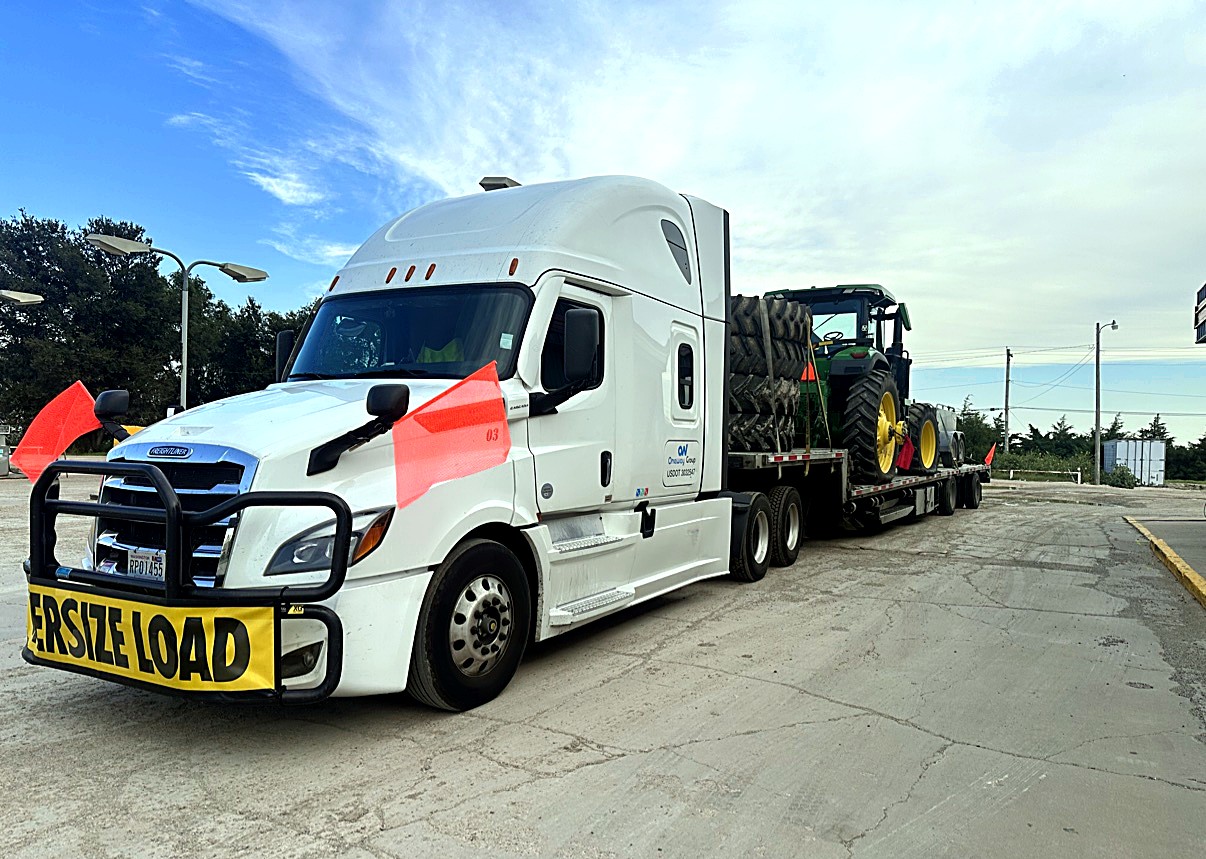 Oneway Group Freightliner with OVERSIZE LOAD banner and orange flags hauling a John Deere tractor and stacked tires on a flatbed