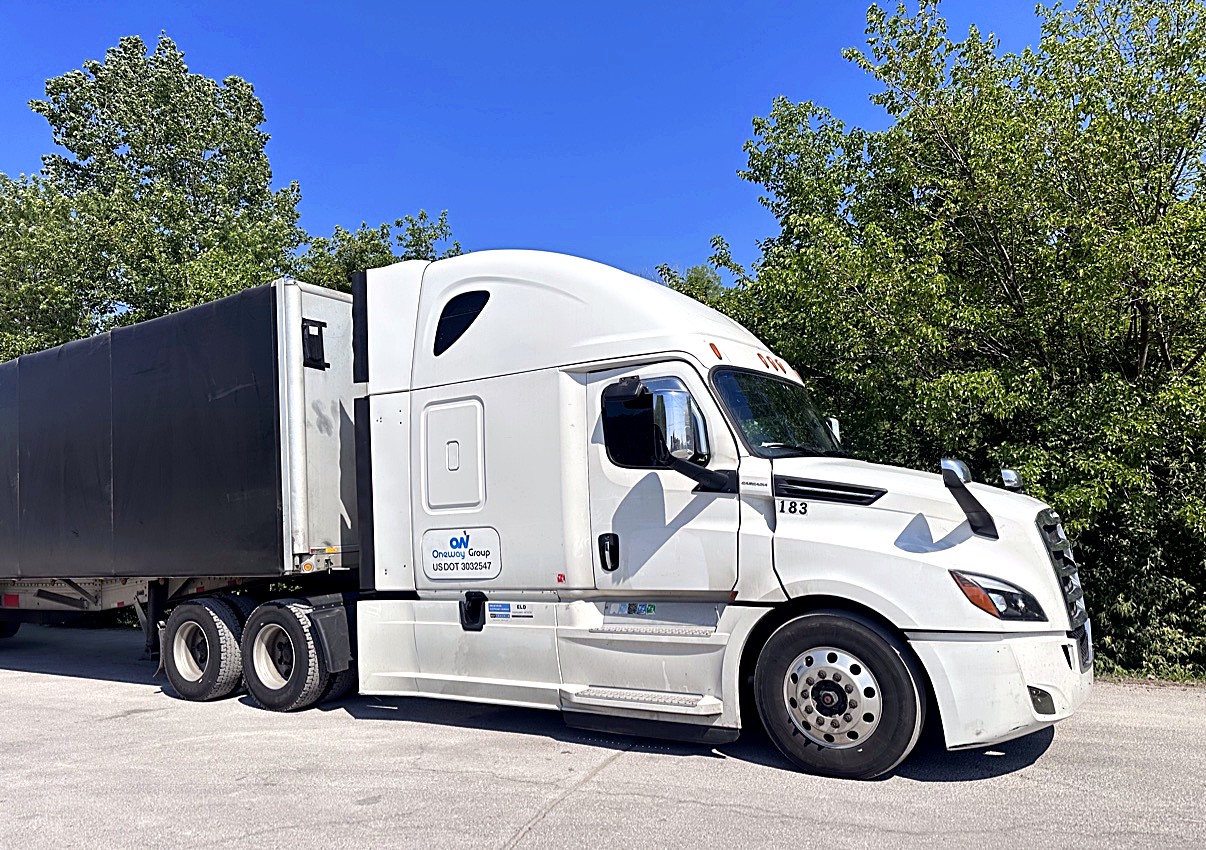 Oneway Group Freightliner Cascadia unit 183 in side profile hooked to a covered trailer, green trees behind